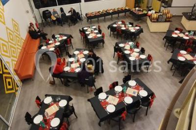 Salle lumineuse avec tables en bois, chaises noires, coin cuisine équipé et grandes fenêtres donnant sur l'extérieur.