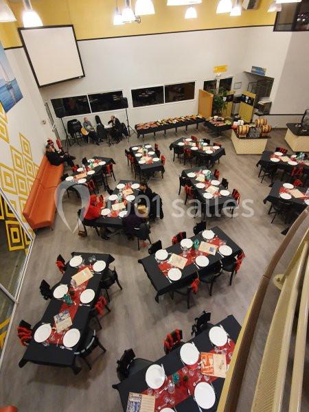 Salle de réception avec tables dressées, nappes noires, serviettes rouges et quelques personnes assises.