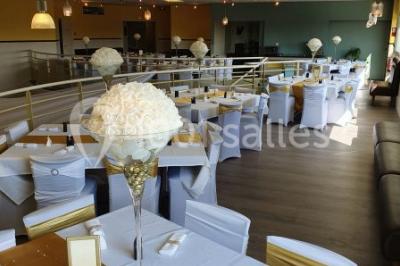 Salle lumineuse avec tables en bois, chaises noires, coin cuisine équipé et grandes fenêtres donnant sur l'extérieur.