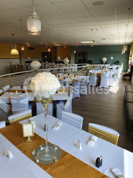 Salle de réception décorée avec des tables alignées, nappes blanches, chemins de table dorés et centres de table fleuris.