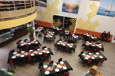 Salle lumineuse avec tables en bois, chaises noires, coin cuisine équipé et grandes fenêtres donnant sur l'extérieur.