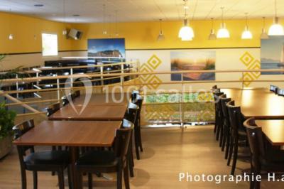 Salle lumineuse avec tables en bois, chaises noires, coin cuisine équipé et grandes fenêtres donnant sur l'extérieur.