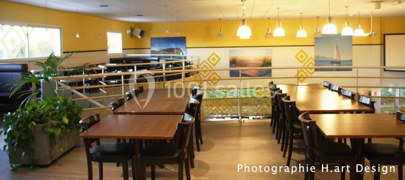 Salle de restaurant lumineuse avec tables en bois, chaises noires, murs jaunes décorés et grandes fenêtres.