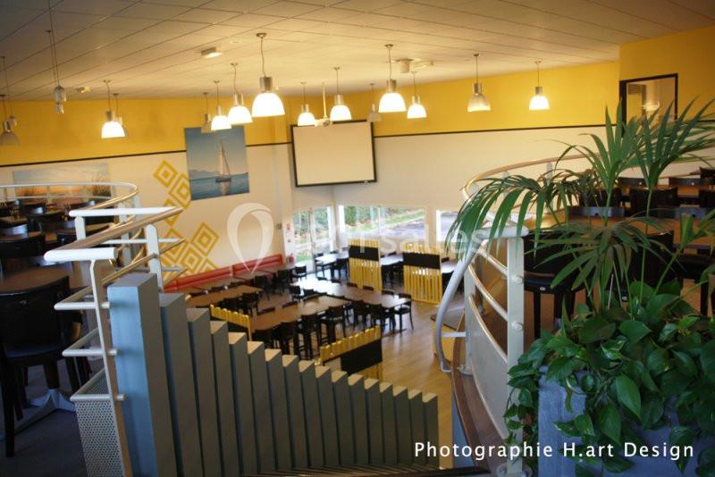 Vue intérieure d'une salle lumineuse avec tables, chaises, plantes et un écran mural, décorée de tons jaunes et blancs.