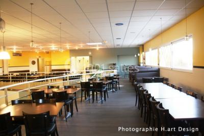 Salle lumineuse avec tables en bois, chaises noires, coin cuisine équipé et grandes fenêtres donnant sur l'extérieur.