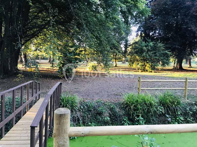 Un petit pont en bois traverse un ruisseau bordé de verdure dans un parc arboré éclairé par une lumière douce.