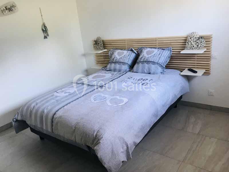 Lit double avec tête de lit en bois, draps gris à motifs de cœurs, dans une chambre lumineuse au sol carrelé.