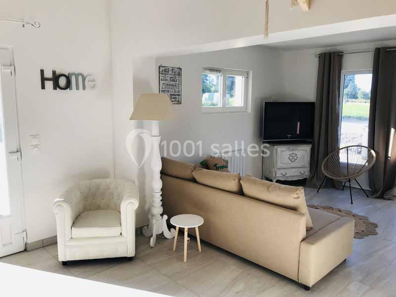 Salon lumineux avec canapé beige, fauteuil blanc, lampe sur pied, table basse et télévision près d'une fenêtre.