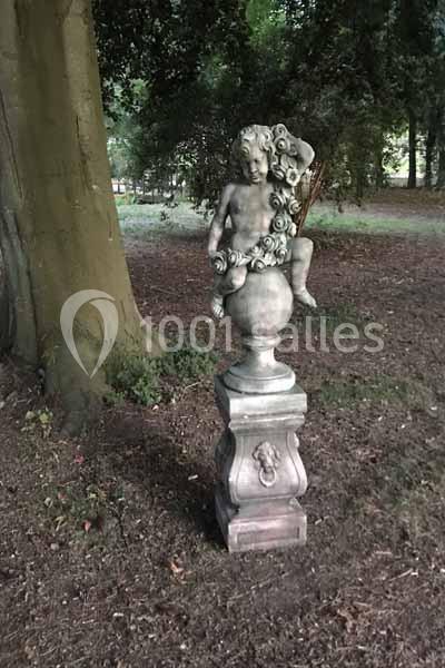 Statue en pierre représentant un enfant assis sur un piédestal, entourée de végétation dans un parc boisé.