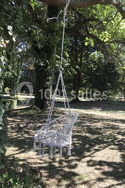 Chaise suspendue en corde blanche accrochée à une branche d'arbre dans un jardin ombragé.