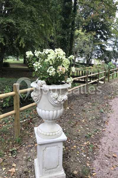 Vase en pierre décoratif avec des fleurs blanches, placé sur un socle près d'une allée bordée de bois et de végétation.