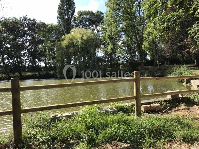 Vue d'un étang entouré d'arbres avec une barrière en bois au premier plan et un ciel partiellement nuageux.