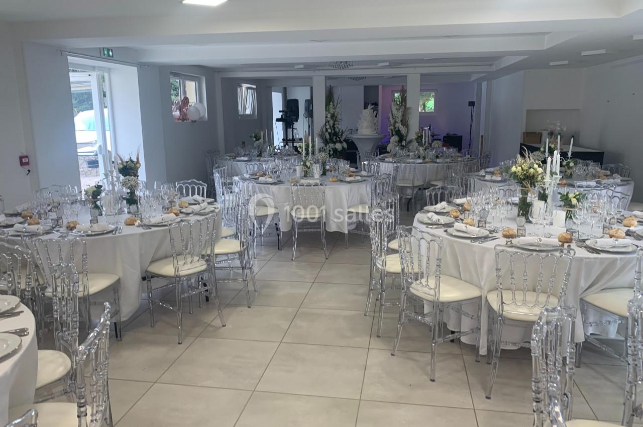 Salle de réception lumineuse avec tables rondes dressées, nappes blanches, chaises transparentes et décorations florales.