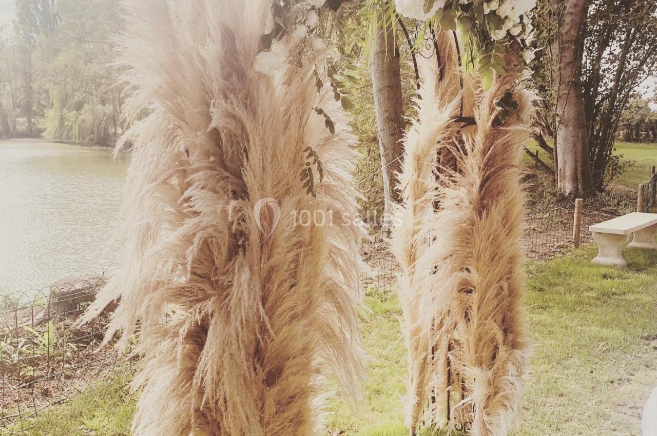 Arche décorée de fleurs blanches et de plumeaux de pampa, installée dans un jardin près d'un étang.
