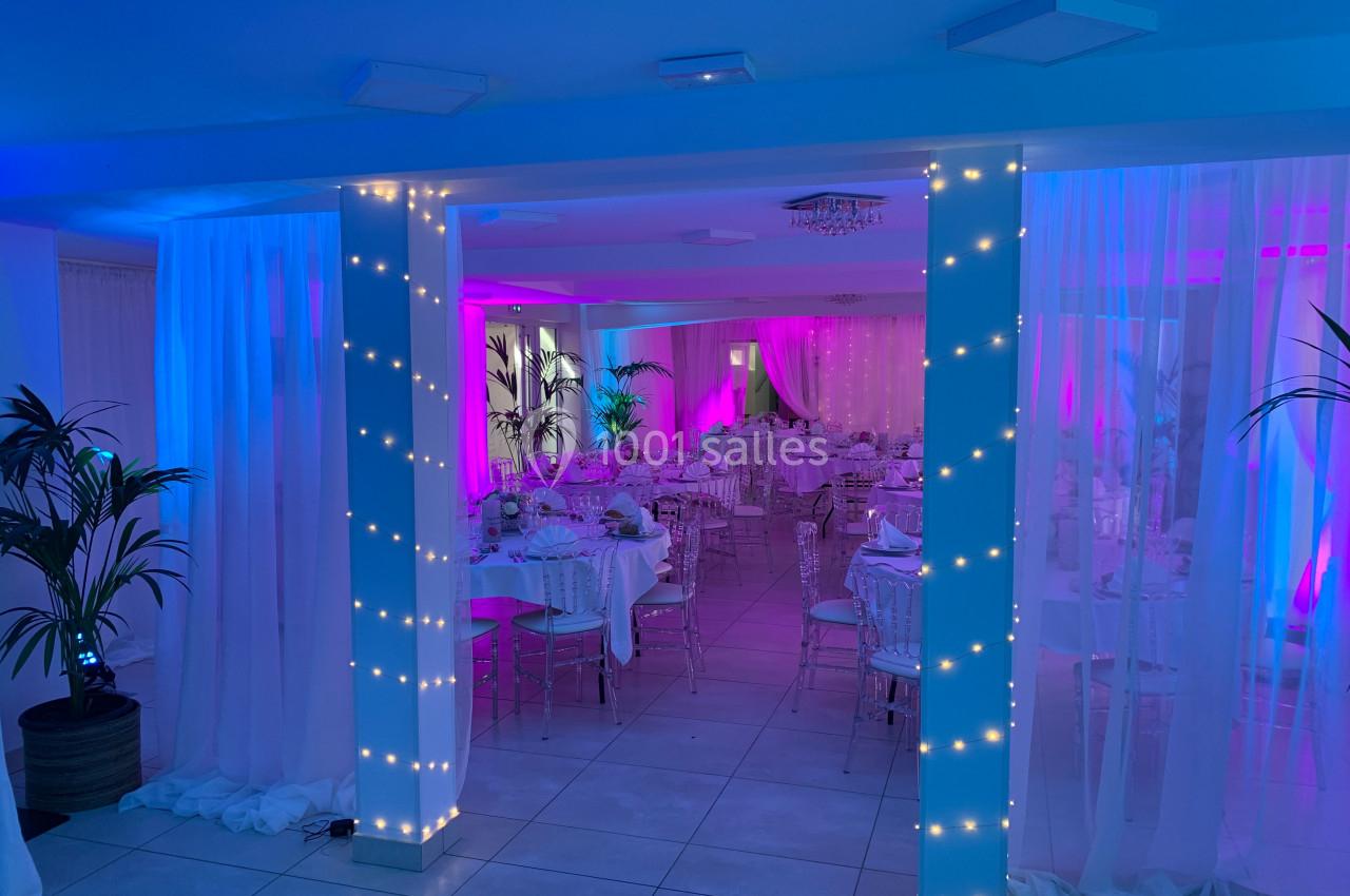 Salle de réception décorée avec des guirlandes lumineuses, des tables dressées et un éclairage rose et bleu.