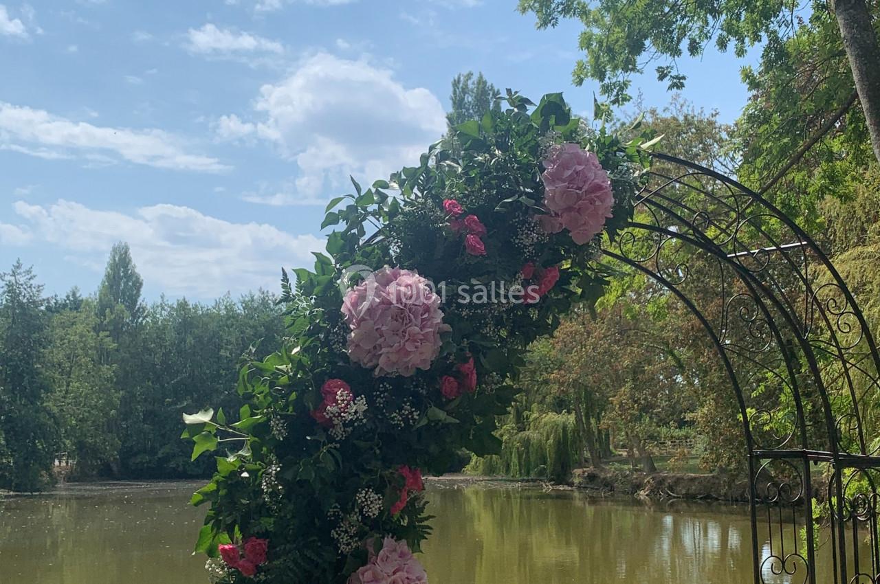 Arche décorée de fleurs roses et feuillage, placée devant un étang entouré d'arbres sous un ciel ensoleillé.