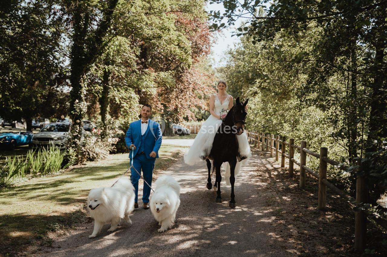 Un homme en costume bleu marche avec deux chiens blancs tandis qu'une femme en robe blanche monte un cheval noir.