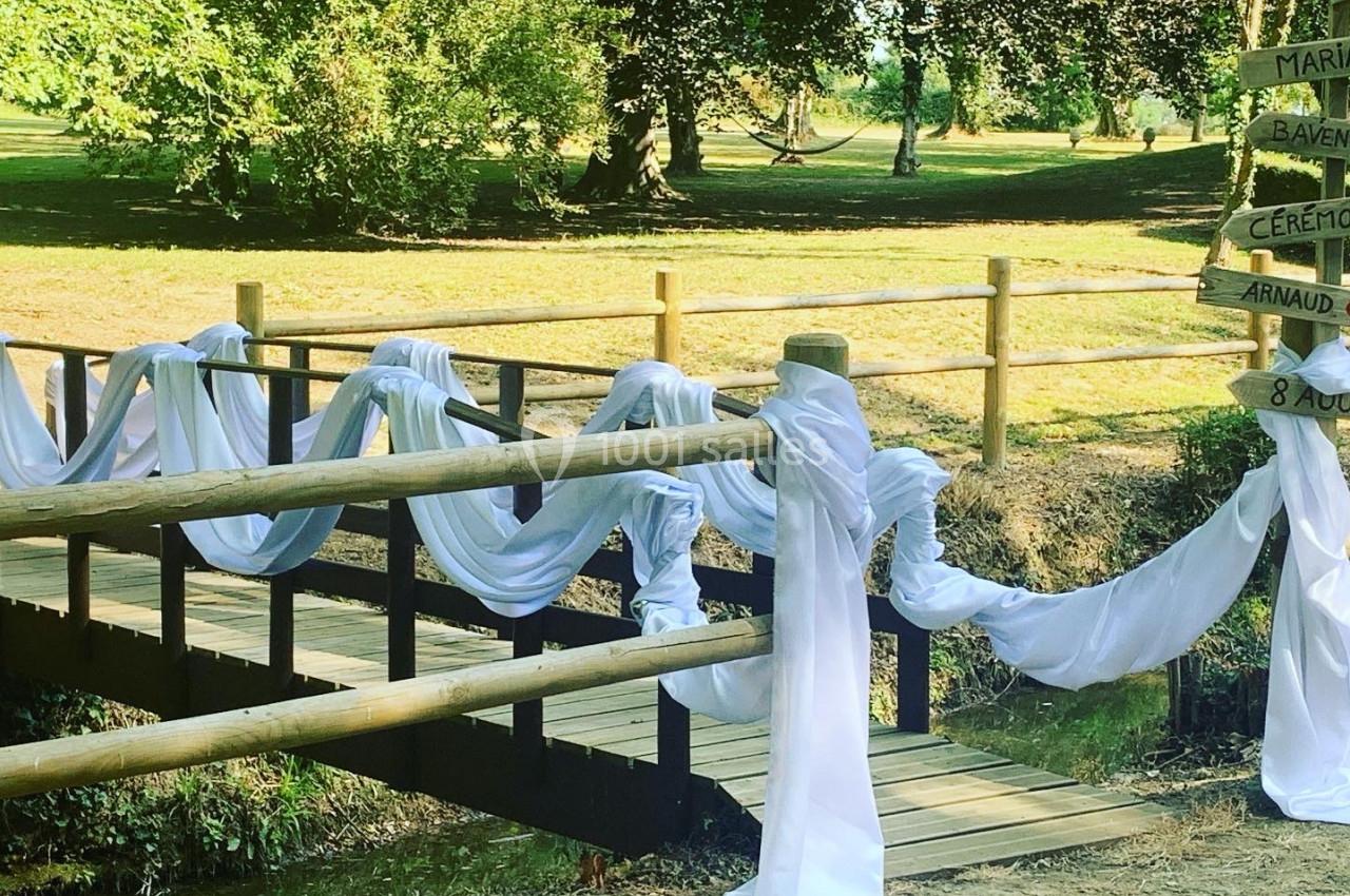 Petit pont en bois décoré de voilages blancs dans un cadre verdoyant, avec un panneau indiquant une cérémonie.