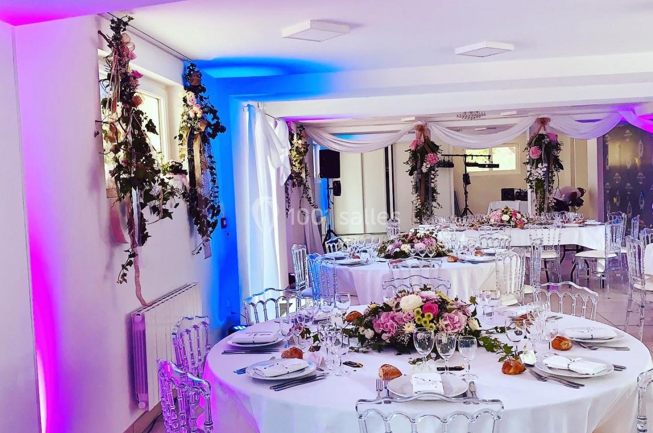 Salle de réception décorée avec des tables rondes, nappes blanches, chaises transparentes et arrangements floraux élégants.