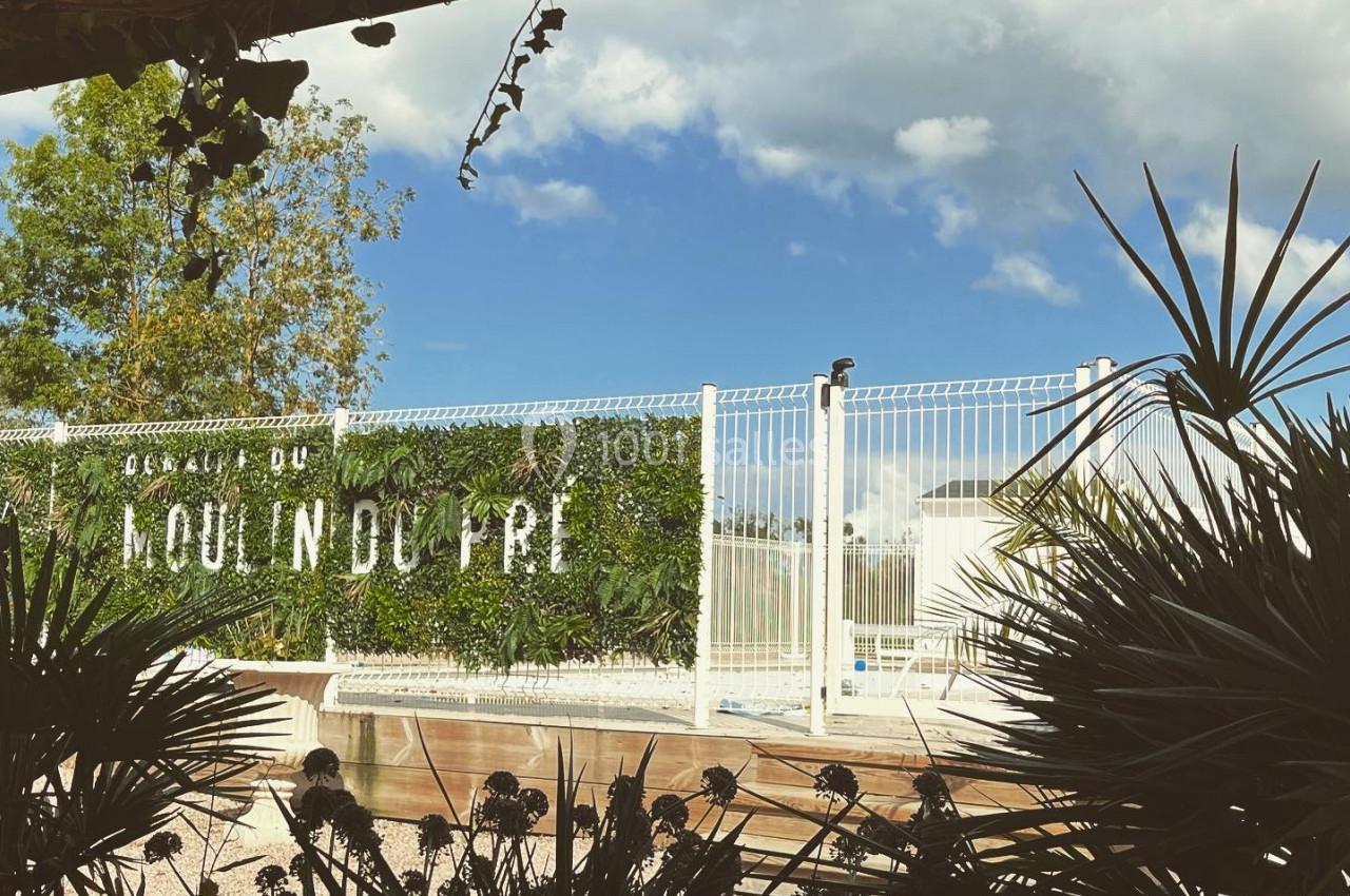 Vue d'un jardin ombragé avec des plantes en premier plan et une clôture blanche devant un ciel partiellement nuageux.