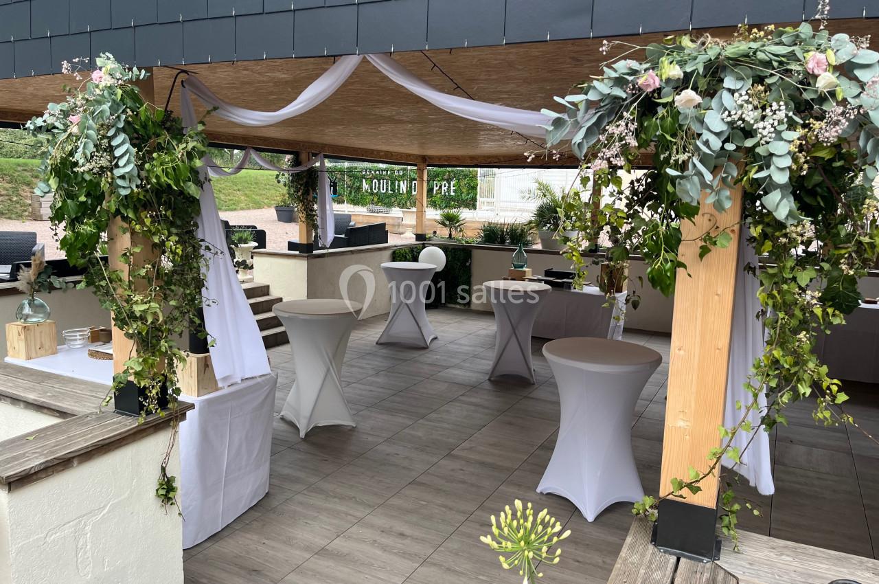 Espace extérieur décoré pour un événement, avec tables hautes, voilages blancs et arrangements floraux suspendus.