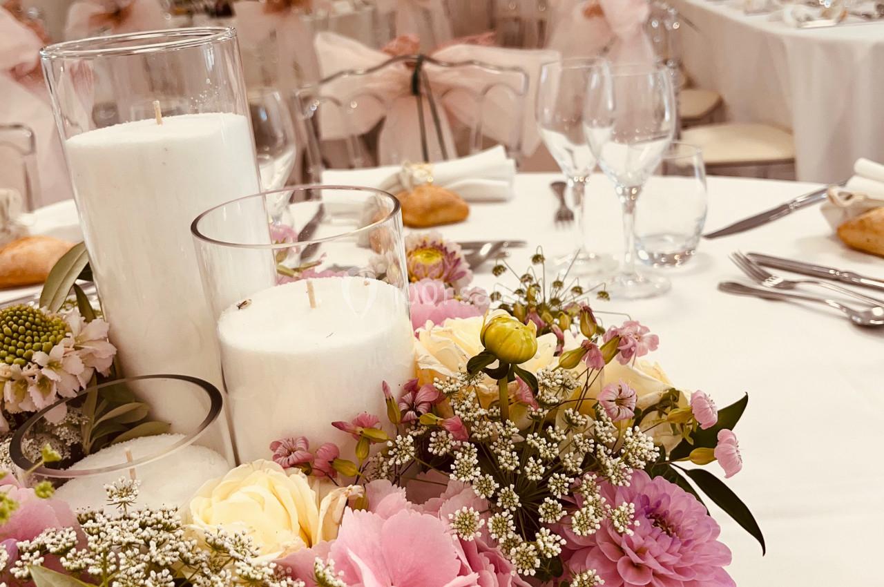 Centre de table floral avec bougies blanches, entouré de vaisselle et nappes blanches dans une salle de réception.