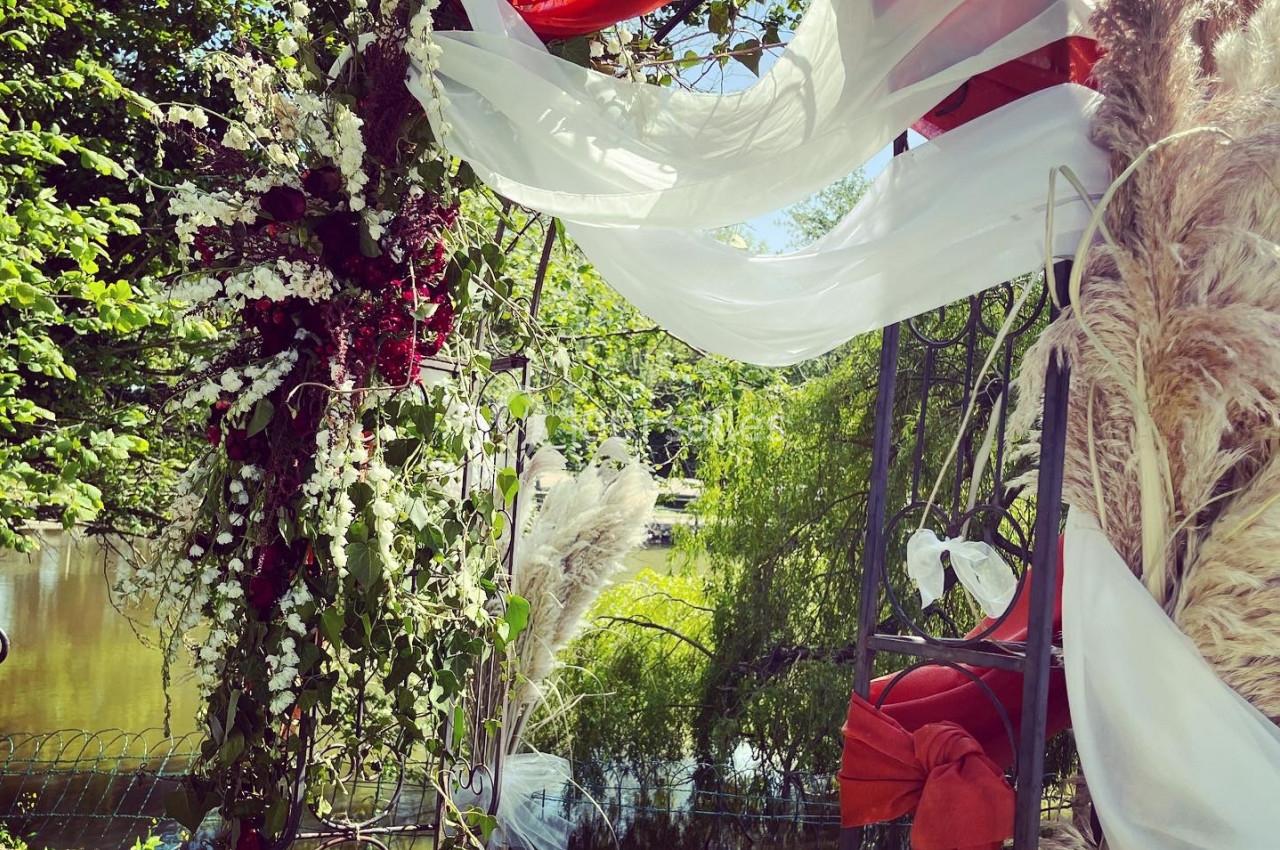 Décoration florale avec voiles blancs et rouges, pampas et verdure, installée près d'un plan d'eau et entourée de végétation.