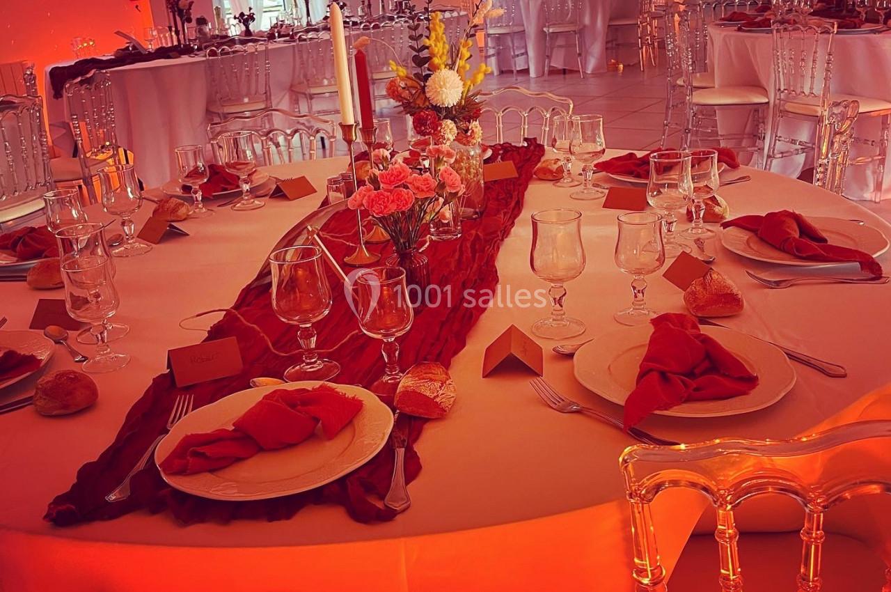 Table ronde décorée pour un événement, avec nappes blanches, serviettes rouges, chandeliers et fleurs au centre.