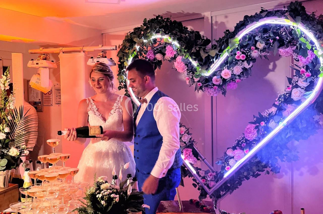 Un couple de mariés coupe un gâteau devant une décoration lumineuse en forme de cœur, entouré de desserts.