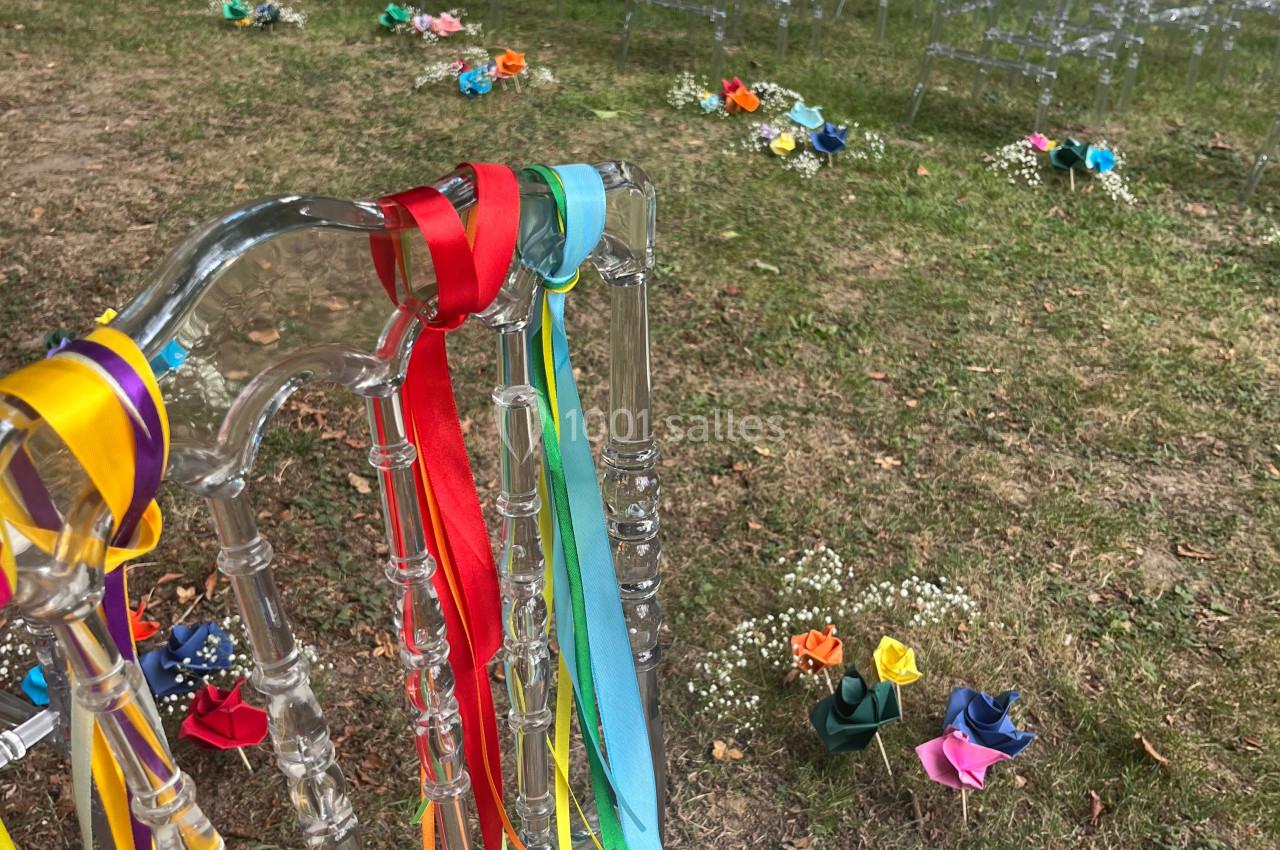 Chaises transparentes décorées de rubans colorés alignées en extérieur, entourées de fleurs disposées sur l'herbe.