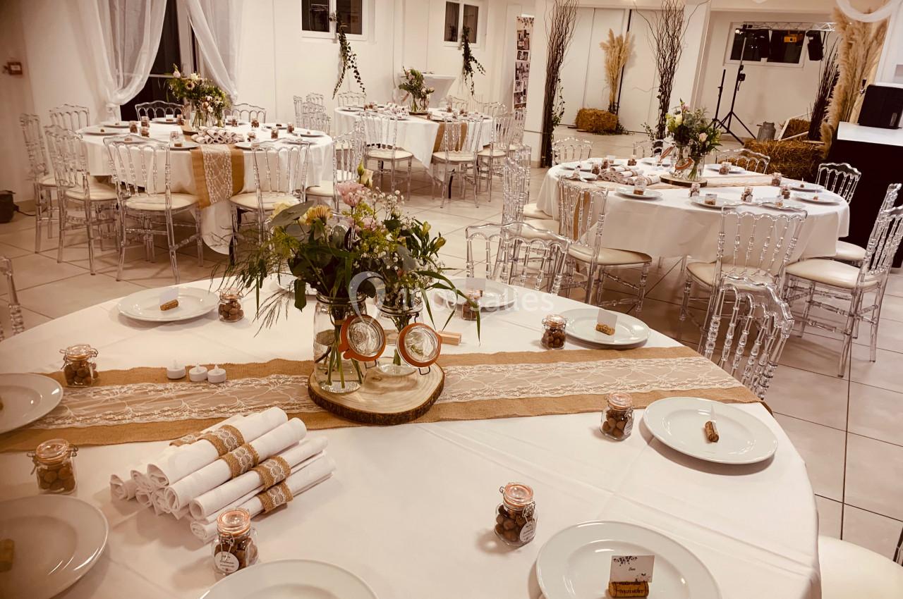 Salle décorée pour un événement, avec tables rondes dressées, nappes blanches, décorations florales et éléments en toile de…