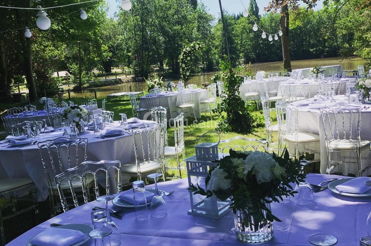 Tables rondes dressées avec nappes blanches et décorations florales dans un jardin ombragé, éclairé par des guirlandes…