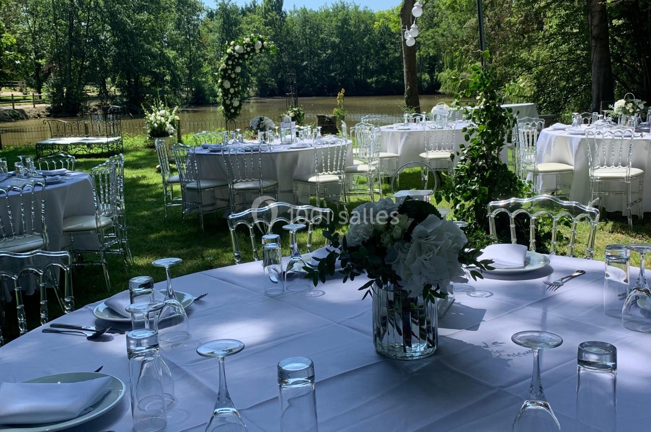 Tables élégamment dressées en extérieur, entourées de verdure et décorées de fleurs blanches sous des guirlandes lumineuses.