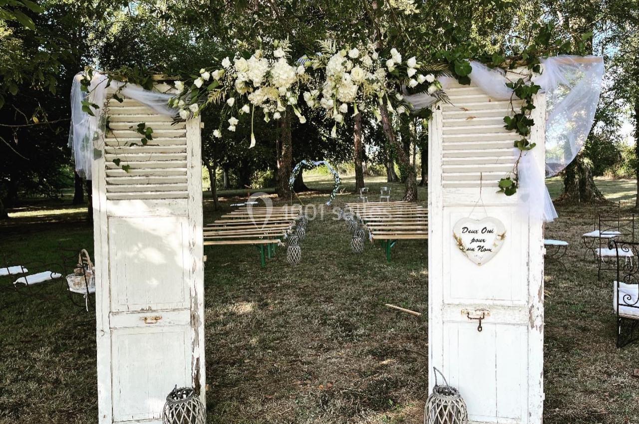 Deux portes blanches anciennes décorées de fleurs blanches et de voilages, encadrant une allée extérieure.
