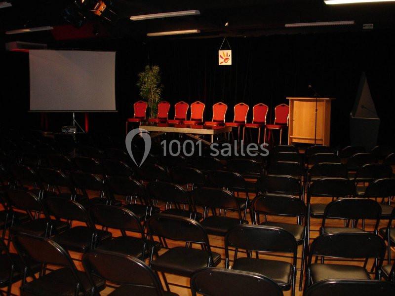 Salle de conférence avec rangées de chaises noires, une estrade équipée de chaises rouges, un pupitre et un écran de…