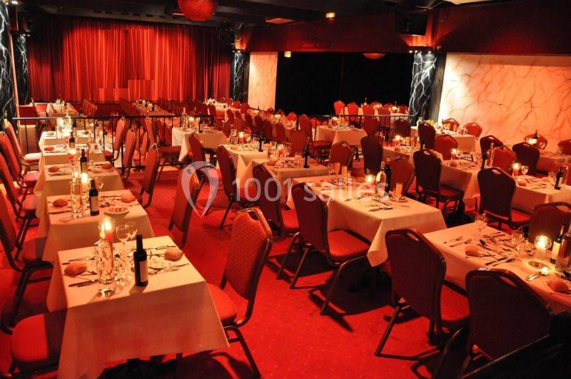 Salle de réception avec des tables dressées, nappes blanches, chaises rouges et éclairage tamisé.