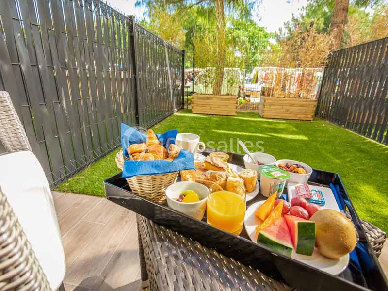 Petit-déjeuner varié avec fruits, viennoiseries et boissons, servi sur une terrasse ensoleillée avec vue sur un jardin.