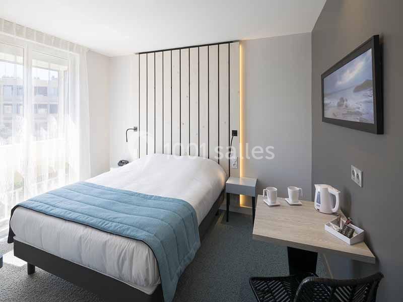 Chambre lumineuse avec lit double, table équipée d'une bouilloire et de tasses, près d'une fenêtre avec rideaux translucides.