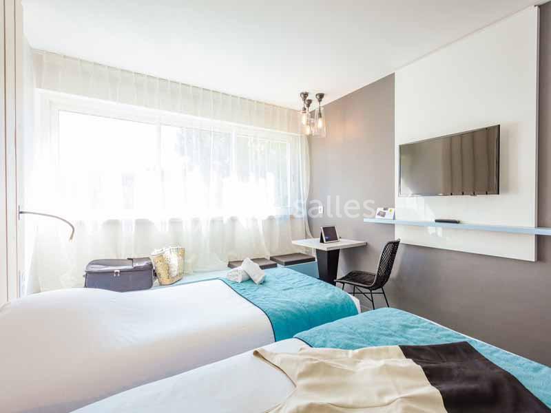 Chambre lumineuse avec deux lits simples, bureau, télévision murale et grande fenêtre avec rideaux translucides.