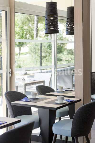 Salle lumineuse avec tables dressées, chaises rembourrées et vue sur un espace extérieur verdoyant à travers de grandes…