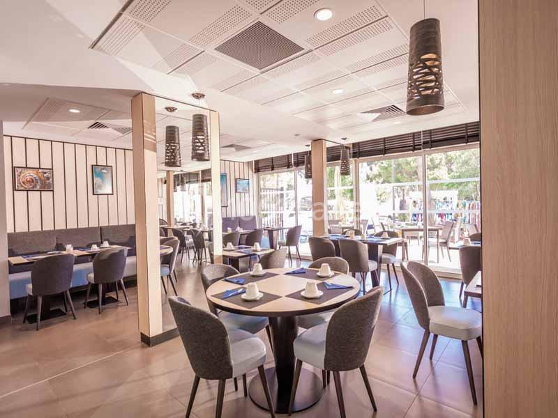 Salle de restaurant lumineuse avec tables dressées, chaises modernes et grandes baies vitrées donnant sur l'extérieur.
