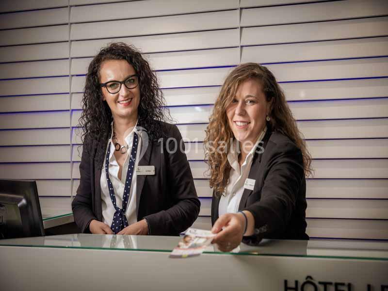 Deux employées souriantes en uniforme accueillent des clients derrière un comptoir d'accueil moderne.