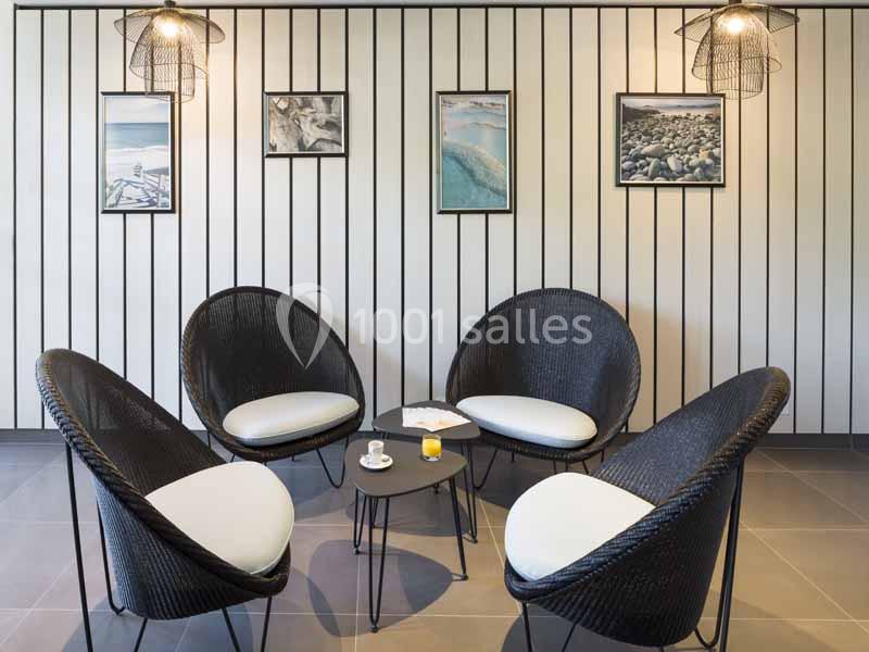 Quatre fauteuils noirs avec coussins clairs entourent une table basse dans une pièce décorée de cadres et luminaires…