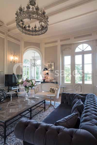 Salon élégant avec moulures, lustres, canapé capitonné, table basse en bois et grandes fenêtres donnant sur un jardin.