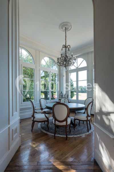 Salle à manger lumineuse avec table ronde, chaises élégantes et grandes fenêtres cintrées donnant sur un extérieur verdoyant.