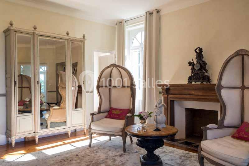 Salon lumineux avec fauteuils capitonnés, table basse en bois, cheminée décorative et armoire à miroir.