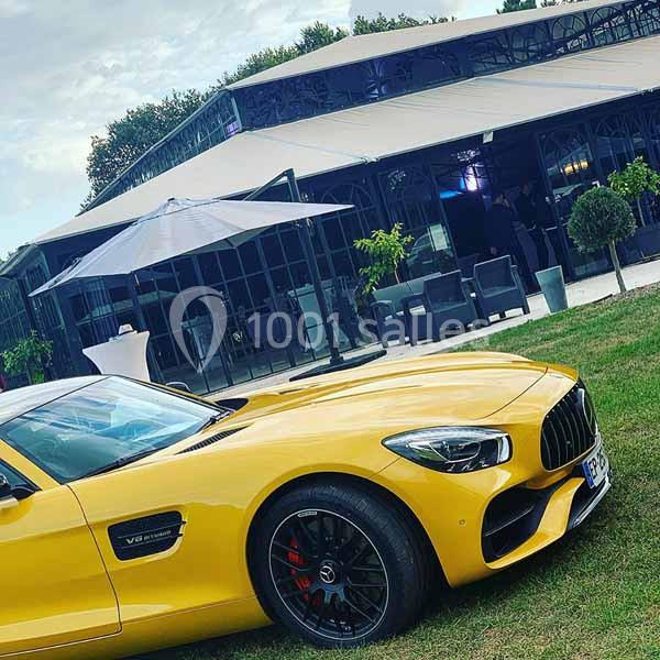 Voiture sportive jaune garée sur une pelouse devant un bâtiment moderne avec terrasse et mobilier extérieur.