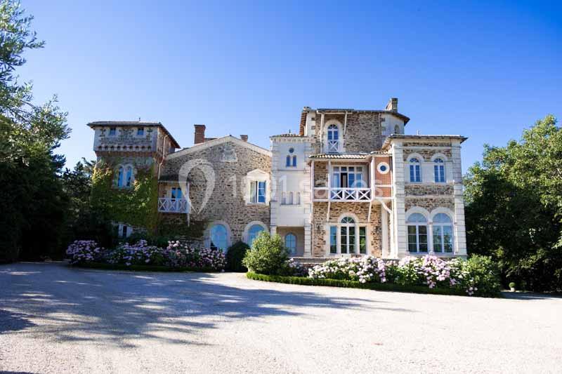 Grande maison en pierre avec des balcons et des fenêtres blanches, entourée de buissons fleuris et d'arbres.