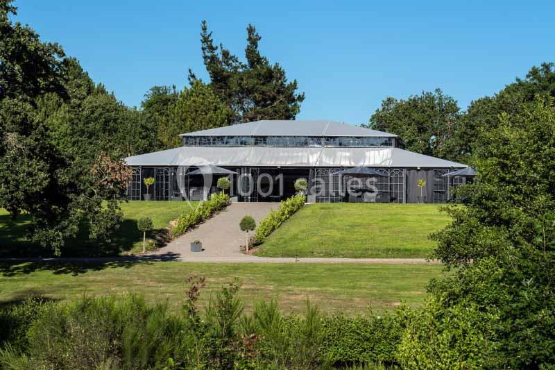 Bâtiment moderne en verre et métal entouré de pelouses et d'arbres sous un ciel bleu.