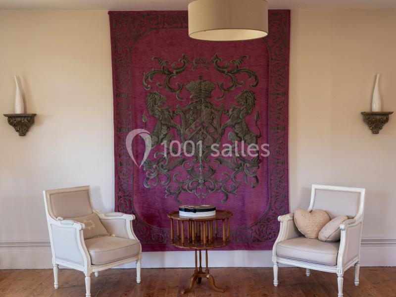 Deux fauteuils blancs entourent une table en bois devant une tapisserie violette ornée d'un motif héraldique.